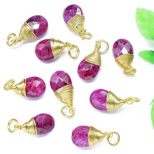 Puede incluir: Diez colgantes de piedra preciosa de color rosa en forma de lágrima con alambre dorado envuelto alrededor. Los colgantes están dispersos sobre una superficie blanca.