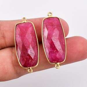 May include: Two rectangular ruby gemstone pendants with gold-toned metal settings and jump rings. The gemstones are faceted and have a deep red colour.