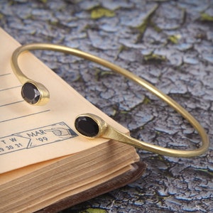 May include: A gold bangle bracelet with two black oval-shaped gemstones. The bracelet is open and rests on a vintage book.