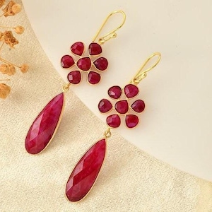 Pode incluir: Brincos dourados com aglomerados de pedras preciosas redondas, vermelho escuro e pingentes em forma de gota. Os brincos têm um design floral e estão sobre um fundo neutro.