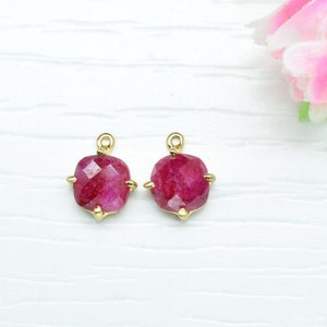 May include: Two ruby-coloured gemstone pendants with gold-coloured settings. The gemstones are square-shaped with faceted surfaces. The pendants are displayed on a white surface, with a pink and green floral accent in the background.