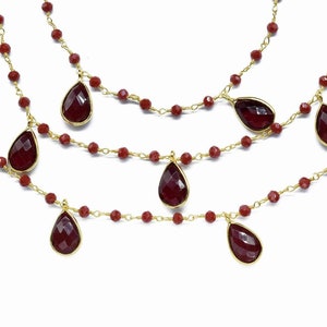 Puede incluir: Un collar de cadena dorada con tres hileras de cuentas rojas y piedras colgantes en forma de lágrima roja.