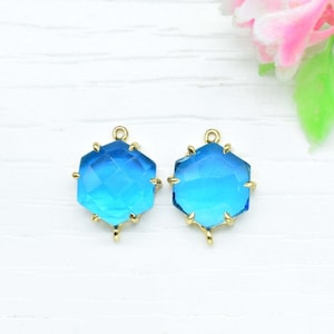 Pode incluir: Dois pingentes de metal dourado com um pingente hexagonal de vidro facetado azul. O vidro tem um degradê de azul escuro para azul claro.