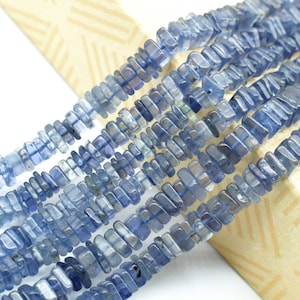 May include: A strand of blue gemstone beads, each bead is a rectangular prism with a slightly irregular shape. The beads are arranged in a row, with the ends of the beads overlapping.