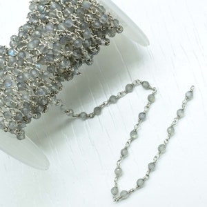 May include: A spool of silver chain with grey gemstone beads. The chain is made of small, round beads that are connected by silver links. The chain is coiled up on the spool, and a small section of the chain is uncoiled and hanging down.