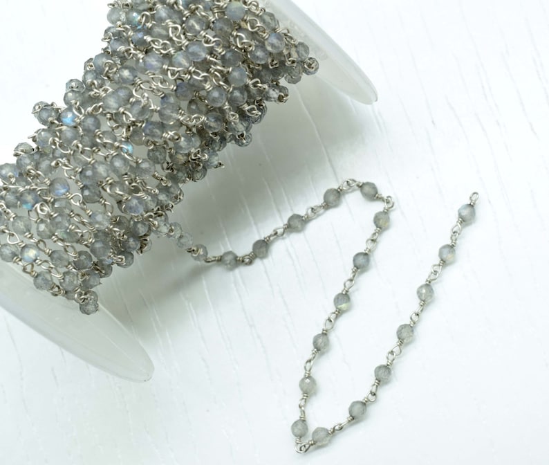 May include: A spool of silver chain with grey gemstone beads. The chain is made of small, round beads that are connected by silver links. The chain is coiled up on the spool, and a small section of the chain is uncoiled and hanging down.