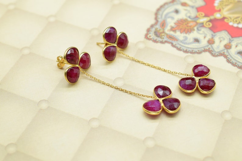 May include: A pair of gold earrings with three ruby-colored stones each, hanging from a delicate gold chain.