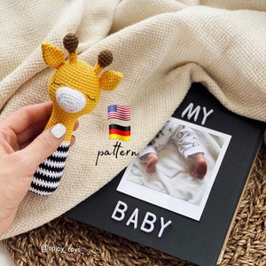 May include: A crocheted giraffe rattle toy with black and white stripes. The toy is held in a hand and is in front of a black photo album with the words "MY BABY" on the front.