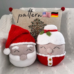 May include: Two handmade Christmas-themed plush ornaments. One is Santa Claus with a red hat and white beard, the other is a Mrs. Claus with a white hat and red dress. They are placed on a white cable-knit blanket.