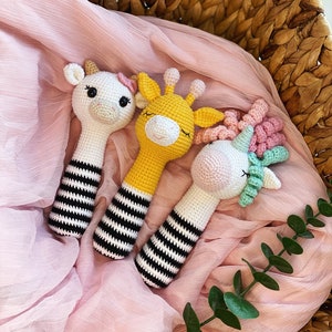 Può includere: Tre giocattoli di peluche all'uncinetto a forma di unicorno, giraffa e mucca. I giocattoli sono tutti bianchi con strisce bianche e nere e hanno occhi neri. L'unicorno ha una criniera e una coda rosa e verde. La giraffa ha un corpo giallo e la mucca ha un corpo bianco. I giocattoli sono sdraiati su uno sfondo di tessuto rosa.