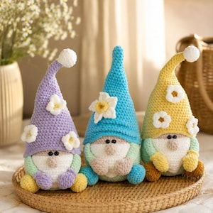 May include: Three handmade crochet gnomes in pastel colours sit on a woven tray. Each gnome has a long, pointed hat with a white pom-pom and a flower accent. The gnomes have white beards, round noses, and small black eyes.
