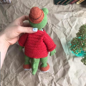Puede incluir: Un cocodrilo de crochet verde que lleva una chaqueta de crochet roja y un sombrero de crochet naranja. El cocodrilo est&aacute; de pie sobre una superficie de papel marr&oacute;n.