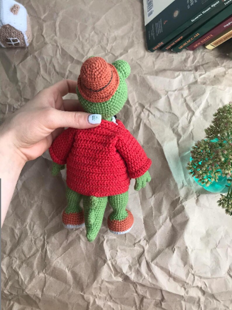 Puede incluir: Un cocodrilo de crochet verde que lleva una chaqueta de crochet roja y un sombrero de crochet naranja. El cocodrilo est&aacute; de pie sobre una superficie de papel marr&oacute;n.