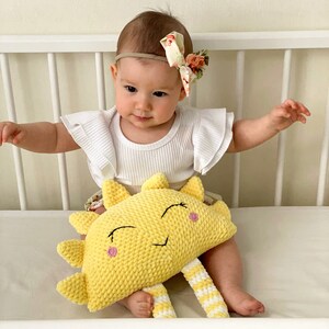 May include: A baby wearing a white shirt with ruffled sleeves and a floral headband is sitting in a crib with a yellow crocheted sun plush toy. The sun has a smiling face with pink cheeks and white and yellow striped legs.