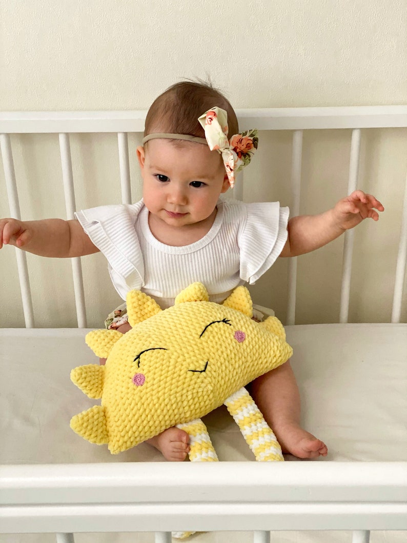 May include: A baby wearing a white shirt with ruffled sleeves and a floral headband is sitting in a crib with a yellow crocheted sun plush toy. The sun has a smiling face with pink cheeks and white and yellow striped legs.