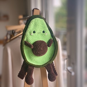 May include: A green crocheted avocado-shaped backpack with brown crocheted legs and a brown crocheted pit. The backpack has a cute cartoon face with eyes and a smile.