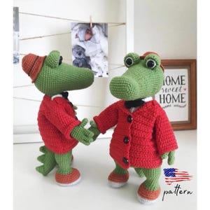 Puede incluir: Dos juguetes de cocodrilo de crochet que llevan chaquetas y sombreros rojos. Los cocodrilos est&aacute;n de pie sobre una superficie blanca con un marco de fotos y un cartel que dice "Home Sweet Home" en el fondo.