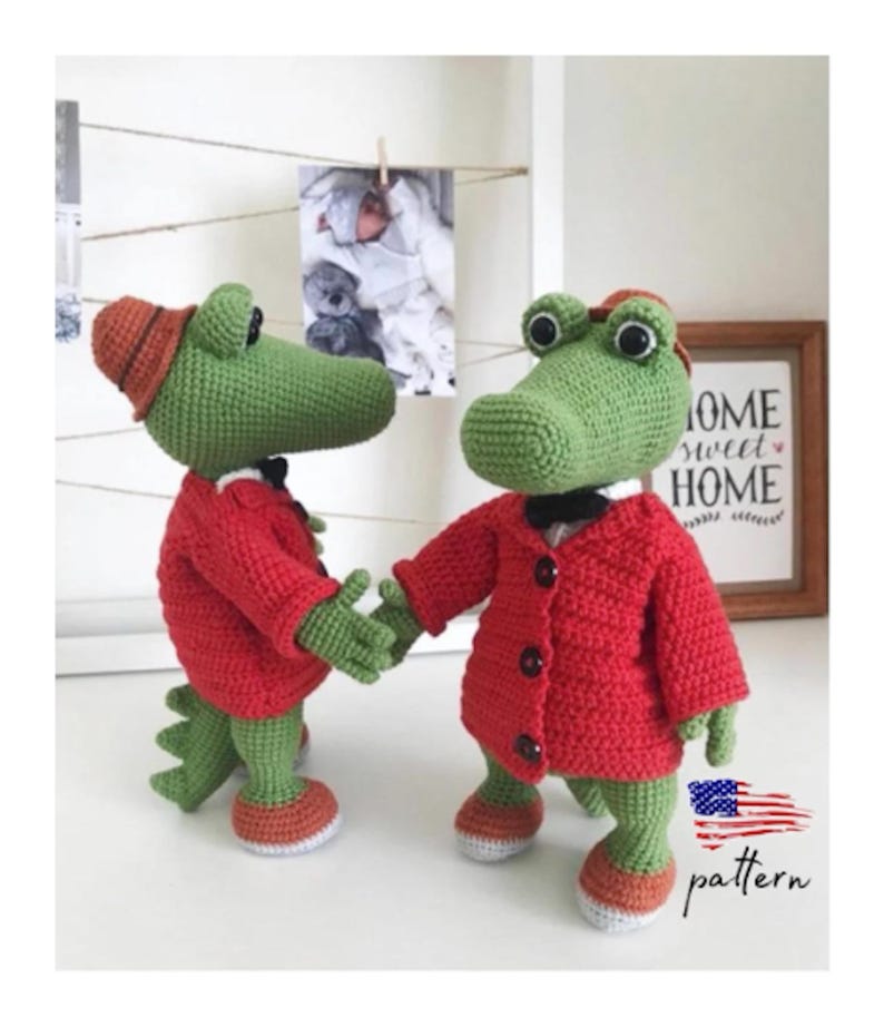Puede incluir: Dos juguetes de cocodrilo de crochet que llevan chaquetas y sombreros rojos. Los cocodrilos est&aacute;n de pie sobre una superficie blanca con un marco de fotos y un cartel que dice "Home Sweet Home" en el fondo.
