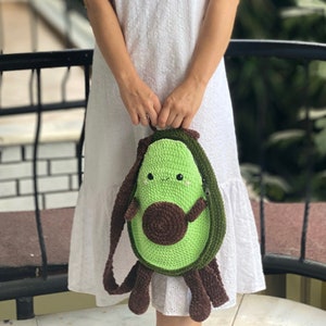 May include: A green crocheted avocado-shaped bag with a brown crocheted pit and a smiling face. The bag has a brown strap and is being held by a person.