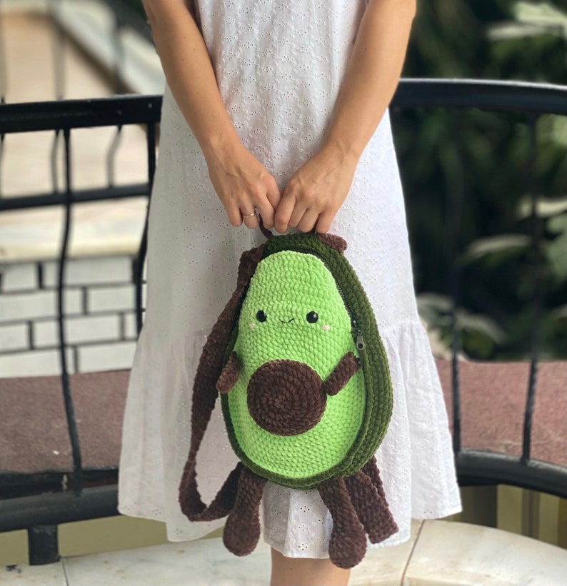 May include: A green crocheted avocado-shaped bag with a brown crocheted pit and a smiling face. The bag has a brown strap and is being held by a person.