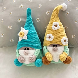 May include: Two handmade crochet gnomes. One gnome has a turquoise hat and a daffodil accent, the other has a yellow hat with white flower accents. Both have white beards, tan noses, and brown feet.