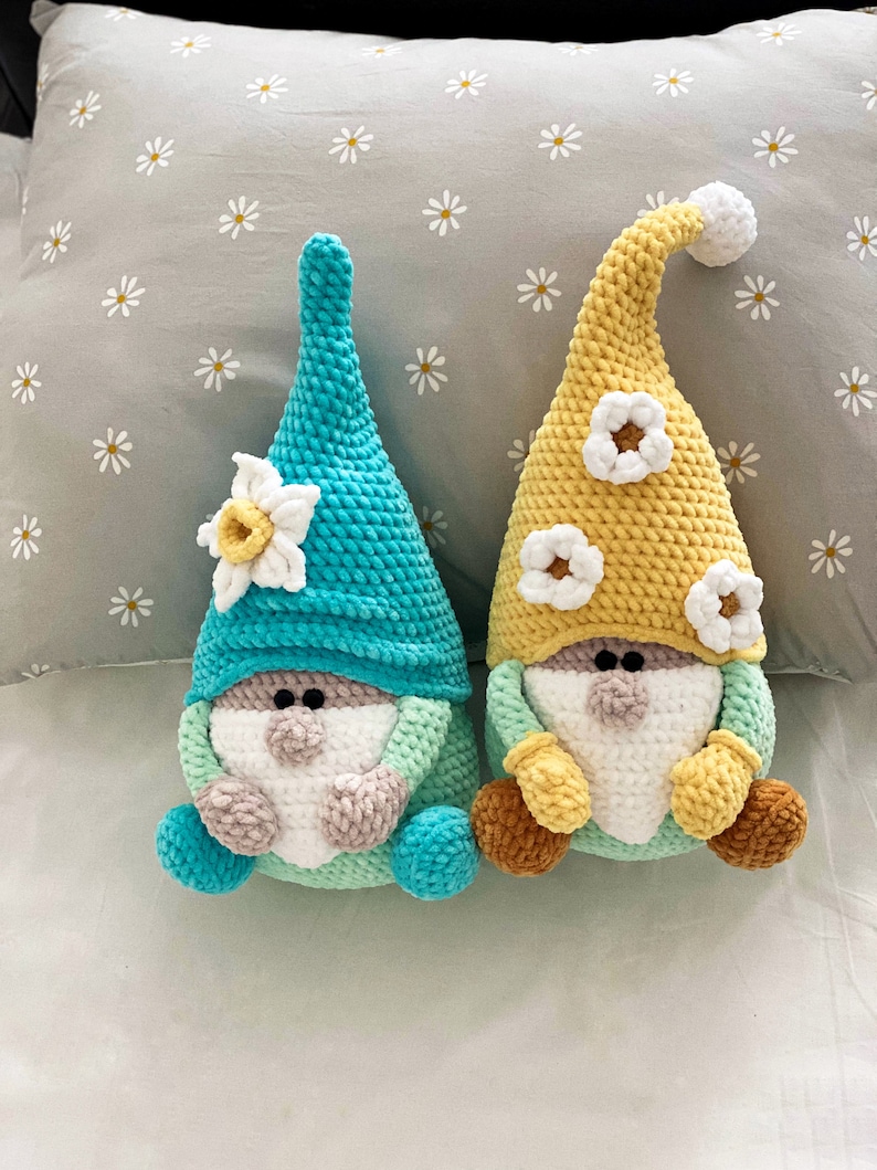 May include: Two handmade crochet gnomes. One gnome has a turquoise hat and a daffodil accent, the other has a yellow hat with white flower accents. Both have white beards, tan noses, and brown feet.