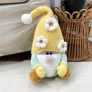 May include: A handmade crochet gnome doll. It features a yellow hat with white flower details, a white beard, a light blue body, and yellow mittens and feet. The doll is placed on a white rug.