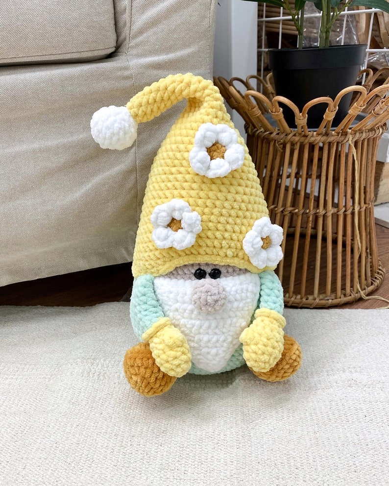 May include: A handmade crochet gnome doll. It features a yellow hat with white flower details, a white beard, a light blue body, and yellow mittens and feet. The doll is placed on a white rug.