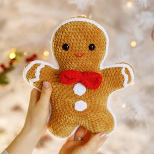 Op de afbeelding: Een pluche peperkoekenman-ornament met een bruin lichaam, witte randen en een rode strik. Het ornament heeft zwarte knoopogen en witte knopen. Het wordt tegen een vage achtergrond gehouden.