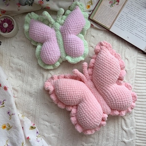 Könnte beinhalten: Zwei handgefertigte Häkel-Schmetterlingskissen. Ein Kissen hat rosa Flügel mit einem grünen Rand, das andere ist ganz rosa. Die Kissen liegen auf einer weißen Zopfstrickdecke, mit einem Stoff mit Blumenmuster im Hintergrund.