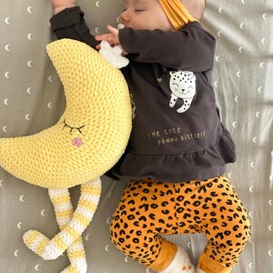 May include: A sleeping baby wearing a mustard yellow headband, a dark gray long-sleeved shirt with a leopard graphic, and orange leopard print pants. A yellow crochet moon toy with striped legs rests beside the baby.