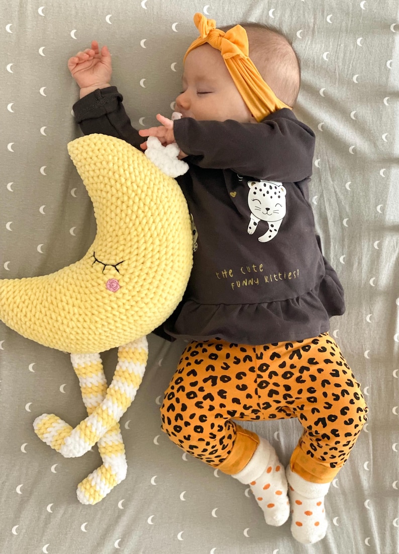 May include: A sleeping baby wearing a mustard yellow headband, a dark gray long-sleeved shirt with a leopard graphic, and orange leopard print pants. A yellow crochet moon toy with striped legs rests beside the baby.