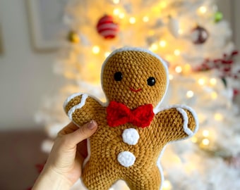 May include: A crocheted gingerbread man toy with a red bow tie and white detailing. The plush is brown with white frosting and black eyes. The background is a blurred Christmas tree with fairy lights.