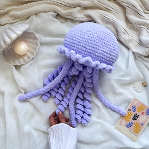 May include: A handmade, lavender-colored crochet jellyfish with a ruffled edge and long, textured tentacles. The jellyfish is resting on a white surface, with a seashell and a small art print nearby. The art print has the text "THE CUT-OUTS".
