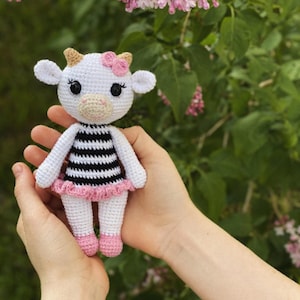 May include: A white crocheted cow doll wearing a black and white striped dress with a pink ruffle. The doll has a pink flower on its head and black eyes.