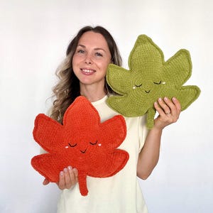 May include: Two leaf-shaped pillows. One is a vibrant orange, the other is a soft green. Both pillows have a smiling face with closed eyes. The pillows are made of a plush, textured material, perfect for home decor.