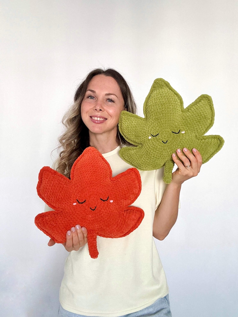 May include: Two leaf-shaped cushions. One is a vibrant orange, the other is a soft green. Both cushions have a smiling face with closed eyes. The cushions are made of a plush, textured material, perfect for home decor.