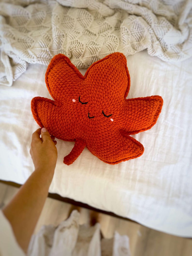 May include: An orange crocheted leaf-shaped pillow with a smiling face. The pillow is on a white bed with a textured blanket. The pillow is approximately 30 cm long.