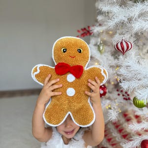 Puede incluir: Un niño sostiene un juguete de peluche de hombre de jengibre frente a su cara. El juguete de peluche es marrón con detalles de glaseado blanco y una corbata de lazo roja. El niño lleva un vestido blanco y sonríe.