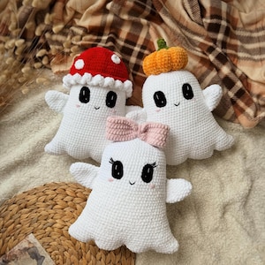 Puede incluir: Tres fantasmas de ganchillo blancos. Un fantasma lleva un sombrero de seta roja con puntos blancos, otro una calabaza naranja en la cabeza y el tercero un lazo rosa claro. Cada fantasma tiene ojos negros, una pequeña sonrisa y mejillas rosadas.