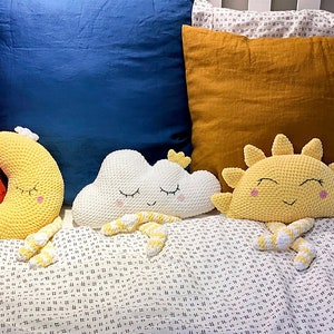 May include: Three crocheted stuffed toys: a yellow crescent moon, a white cloud, and a yellow sun. Each toy has a smiling face with closed eyes and is lying on a white and black patterned bedspread.