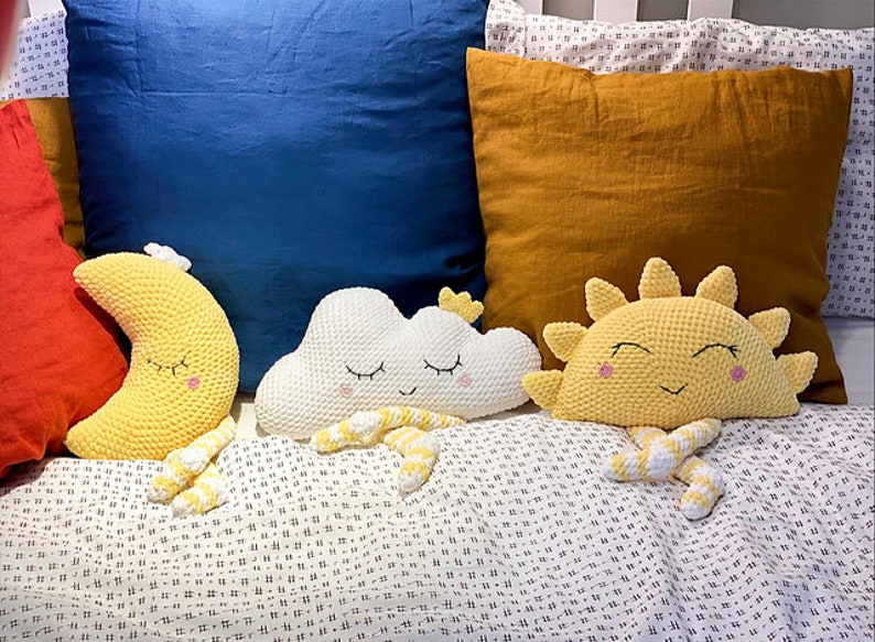 May include: Three crocheted stuffed toys: a yellow crescent moon, a white cloud, and a yellow sun. Each toy has a smiling face with closed eyes and is lying on a white and black patterned bedspread.