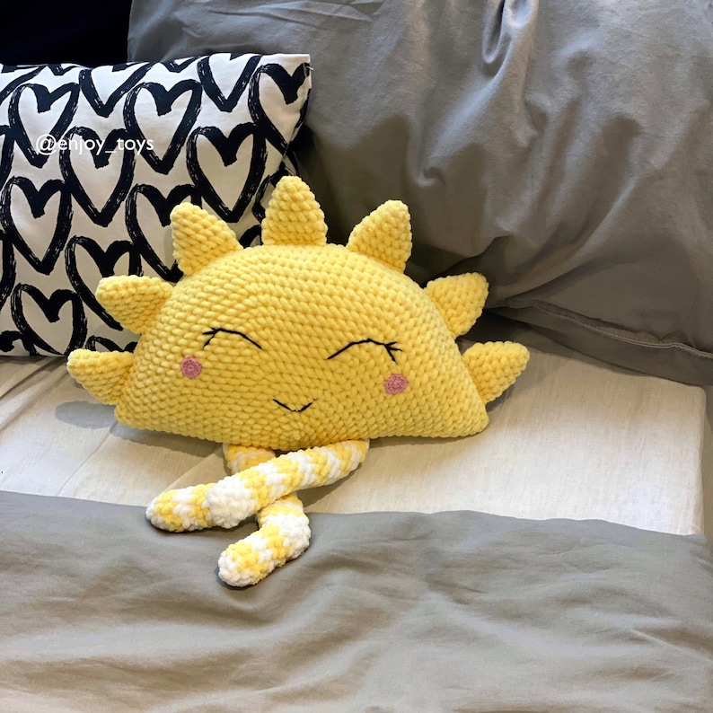 May include: A yellow crocheted sun pillow with a smiling face and pink cheeks. The pillow has white and yellow crocheted legs.