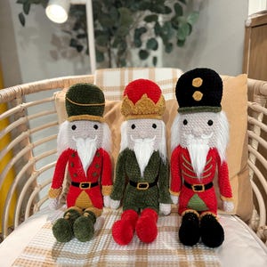 May include: Three handmade crochet nutcracker dolls. Each doll has a white beard, a red or green jacket, and a black belt. The dolls have different colored hats: green, red, and black. The dolls are sitting on a wicker chair with a plaid blanket.