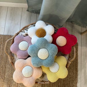 Puede incluir: Una colección de seis cojines de ganchillo con forma de flor en una cesta de mimbre. Los cojines presentan hilo suave y texturizado en blanco, rosa empolvado, azul claro, rojo brillante, melocotón y amarillo, con un centro circular. Texto: @enjoy_toys.