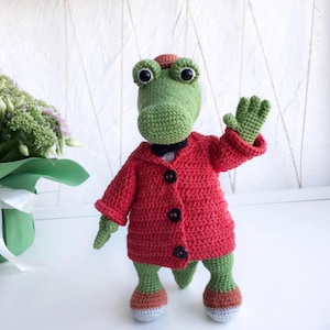 Puede incluir: Un cocodrilo de crochet verde que lleva un abrigo de crochet rojo con botones negros y una pajarita negra. El cocodrilo est&aacute; de pie sobre una superficie blanca.