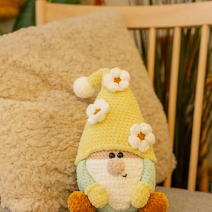 May include: A handmade crochet gnome doll. It features a yellow hat decorated with white flower accents. The gnome has a light green and pink face, a tan nose, and yellow feet. The doll is sitting on a grey surface.