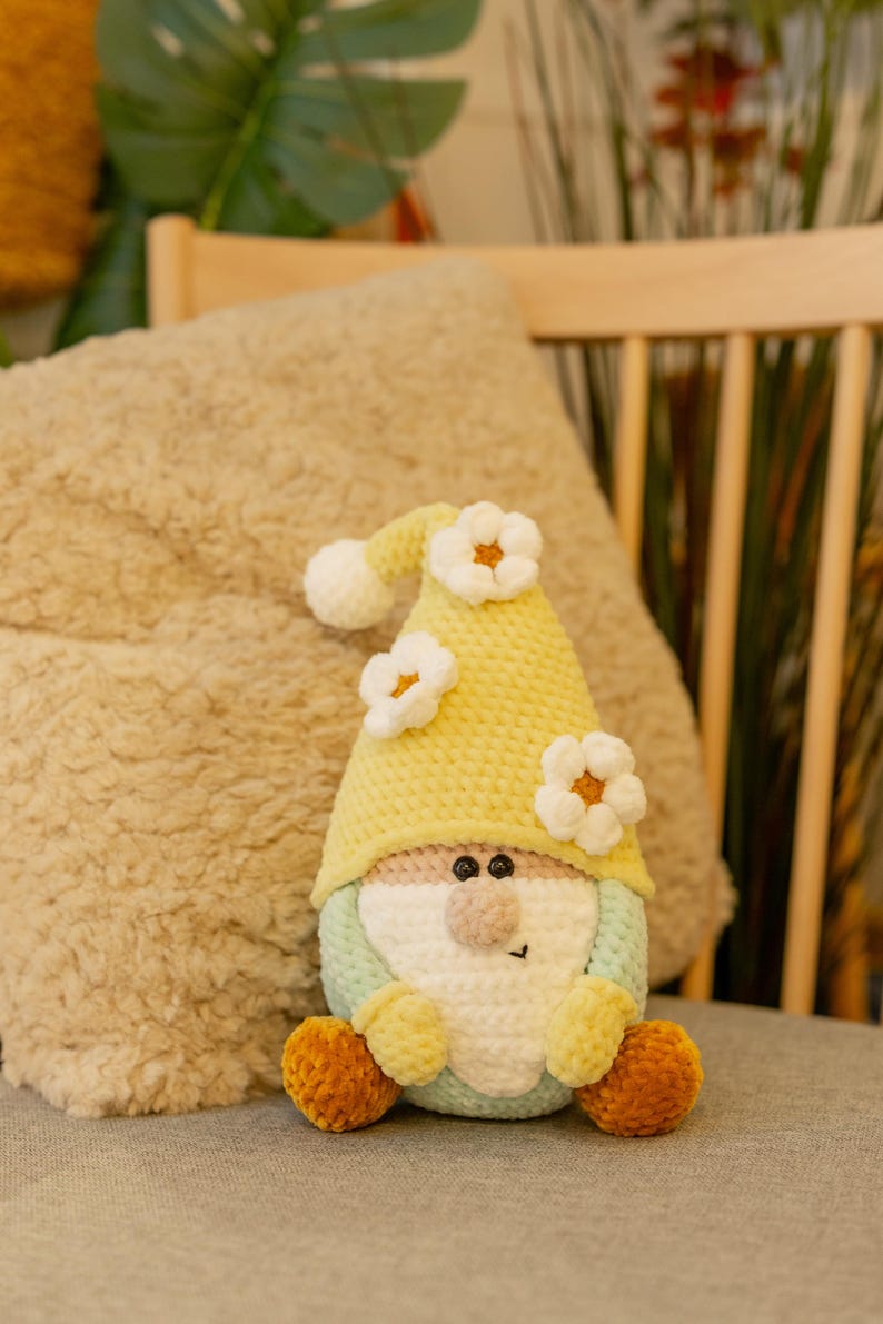 May include: A handmade crochet gnome doll. It features a yellow hat decorated with white flower accents. The gnome has a light green and pink face, a tan nose, and yellow feet. The doll is sitting on a grey surface.