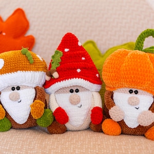 Puede incluir: Tres muñecos de gnomos hechos a mano con ganchillo con sombreros de champiñones y calabazas. Los gnomos tienen barbas blancas, caras sonrientes y cuerpos marrones. Los sombreros son naranjas, rojos y blancos. Los muñecos están sobre una superficie beige.