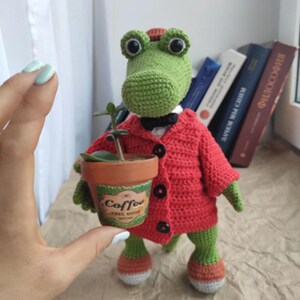 Puede incluir: Un cocodrilo de crochet verde que lleva un c&aacute;rdigan rojo y una pajarita negra sostiene una peque&ntilde;a maceta de terracota con una planta verde y una etiqueta que dice "Coffee Feel Good".
